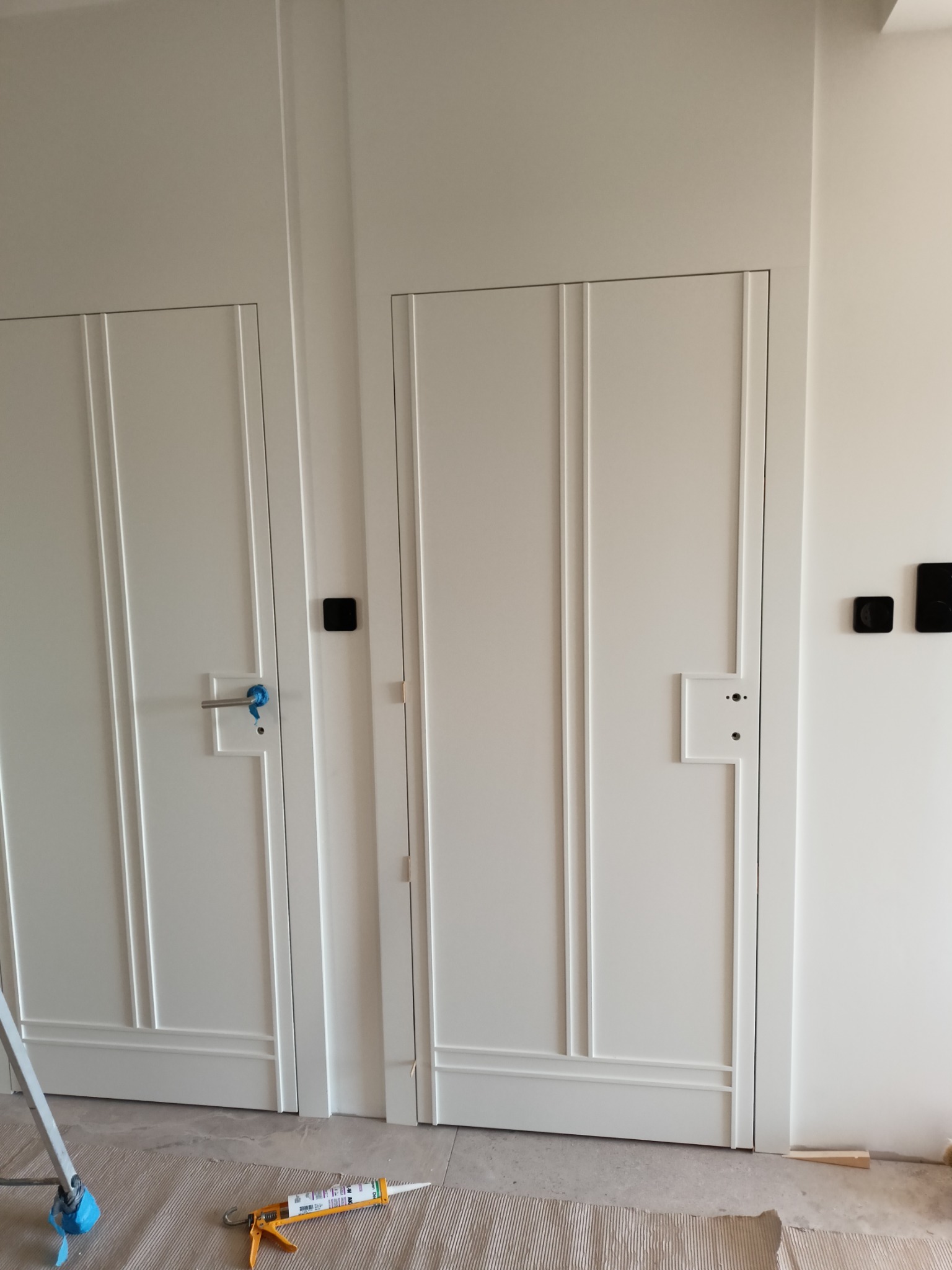 Dwa białe, panelowe drzwi wewnętrzne po montażu, widoczne narzędzia i materiały uszczelniające na podłodze, czujniki na ścianie.