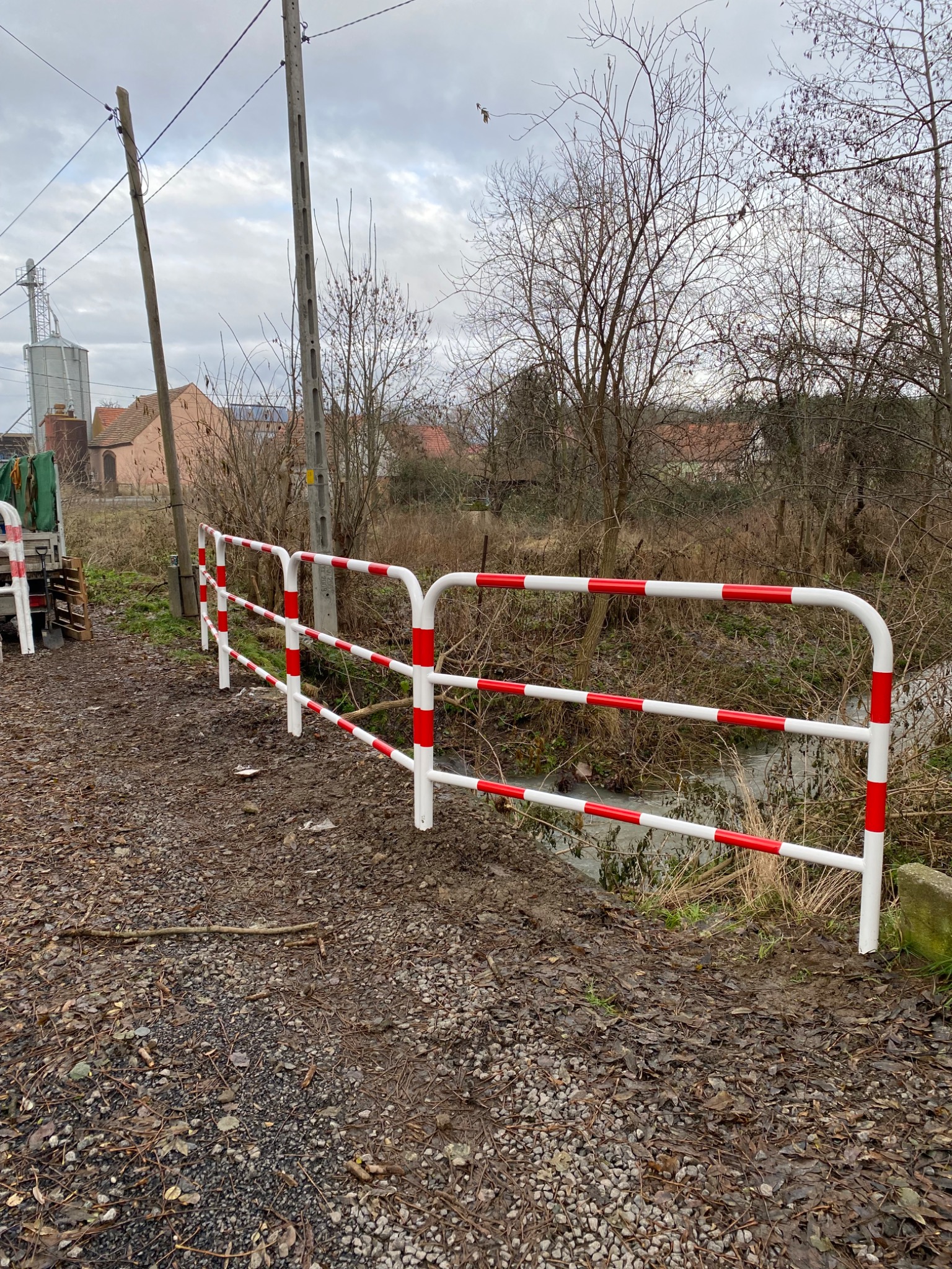 Nowo zainstalowana, biało-czerwona bariera ochronna przy drodze gruntowej, zabezpieczająca przed spadkiem do niewielkiego cieku wodnego; w tle zabudowania gospodarcze i drzewa bezlistne.