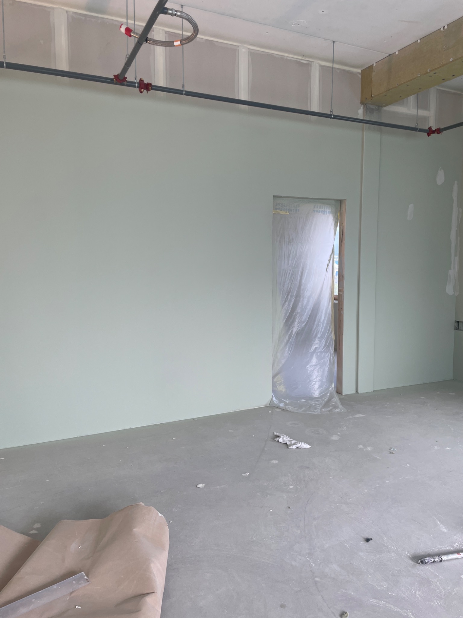 Wnętrze w trakcie wykańczania: ściany pomalowane na jasnozielony kolor, zabezpieczone folią drzwi, widoczne instalacje pod sufitem, na podłodze resztki materiałów budowlanych i narzędzia.