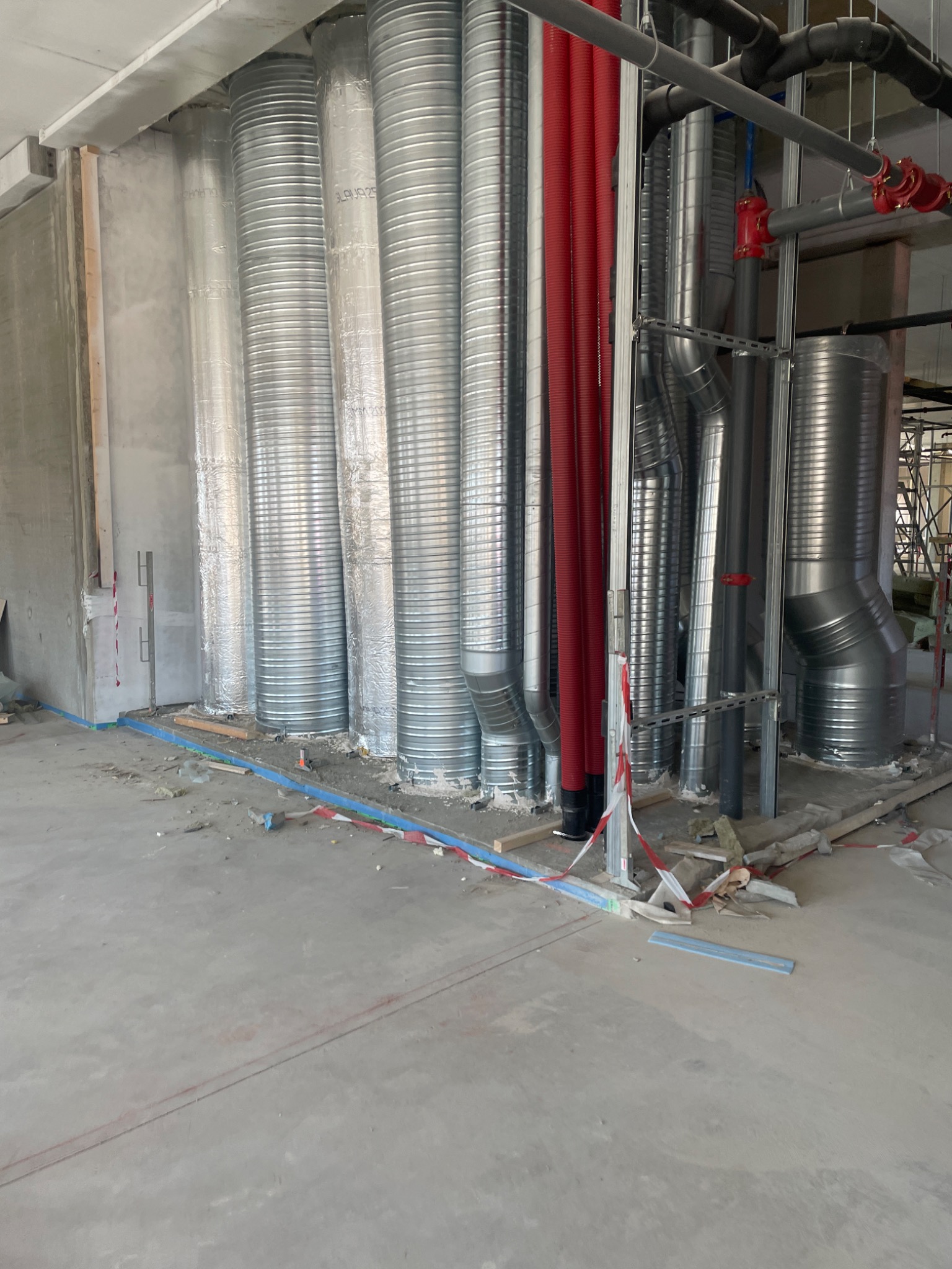 Wnętrze w trakcie prac budowlanych: widoczne surowe ściany, liczne metalowe rury wentylacyjne owinięte folią, czerwone rury instalacyjne, oraz nieporządek na betonowej podłodze z fragmentami...