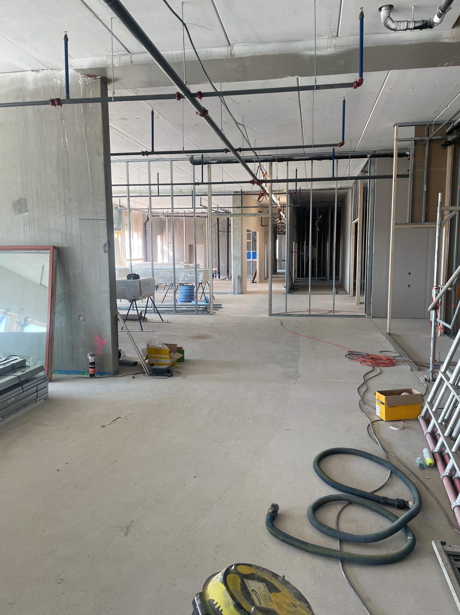 Wnętrze w stanie surowym z widocznymi instalacjami pod sufitem, metalową konstrukcją ścian działowych, betonowymi słupami i przygotowanymi materiałami budowlanymi, sprzętem budowlanym...