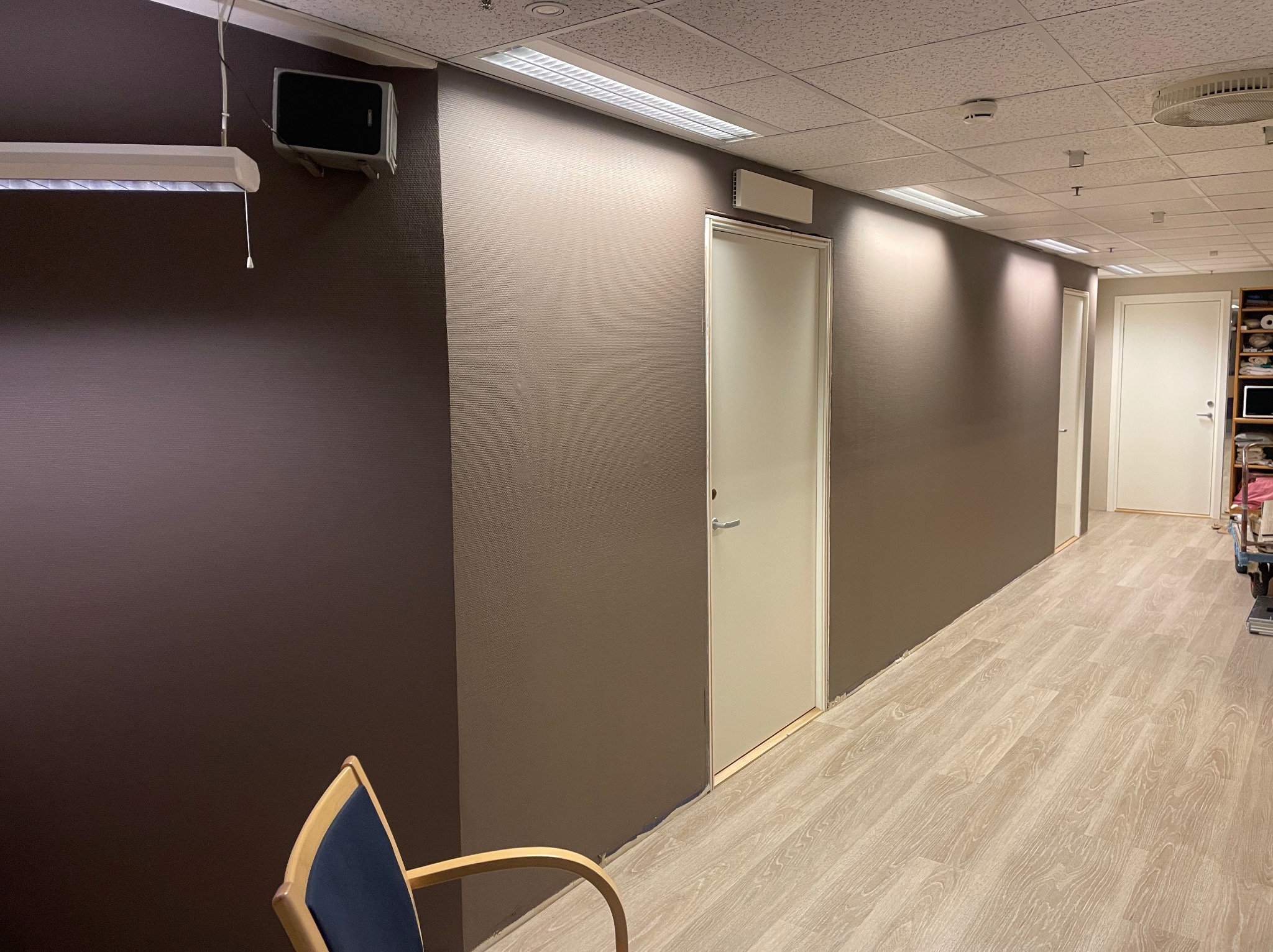 Korytarz biurowy z brązowymi ścianami o fakturze, jasną podłogą imitującą drewno, białymi drzwiami i widocznym krzesłem z niebieskim obiciem w rogu kadru.