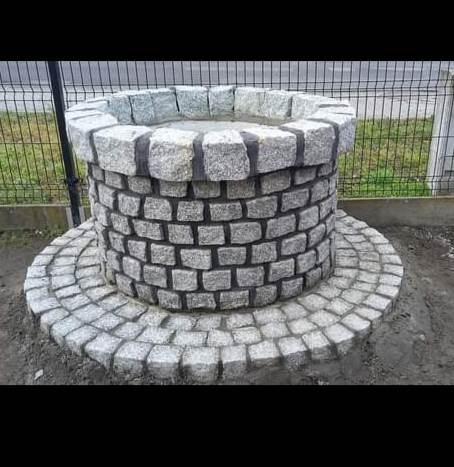 Studnia z szarej kostki granitowej na okrągłej podstawie, widoczna siatka ogrodzeniowa w tle.