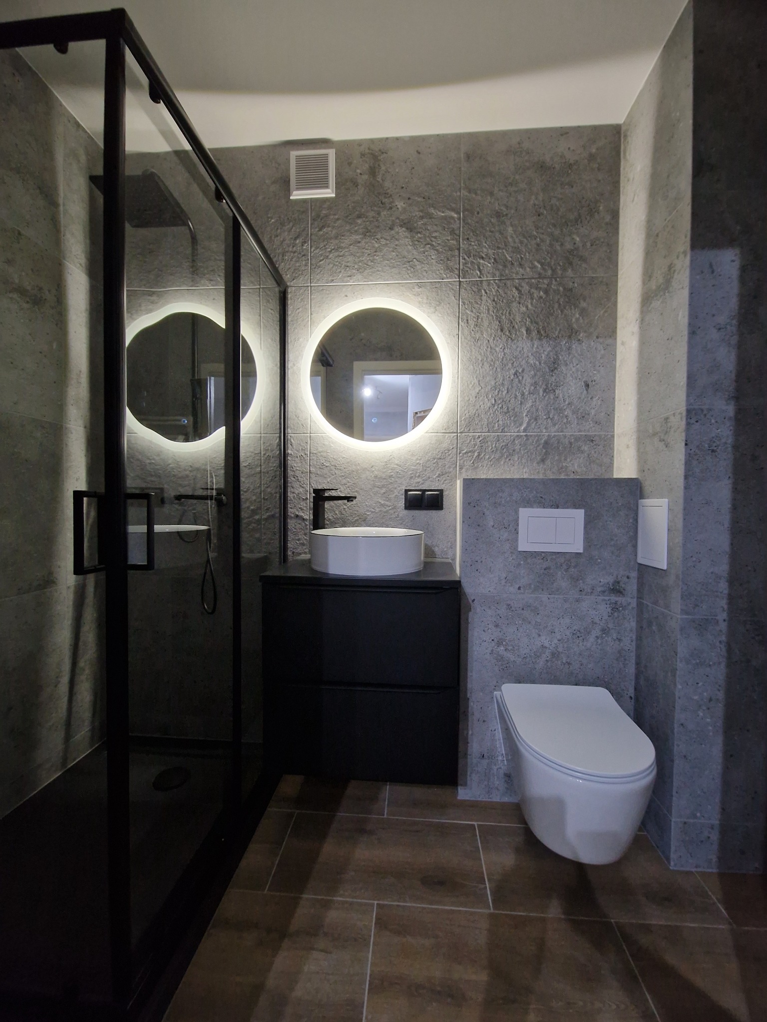 Nowoczesna łazienka z szarymi płytkami imitującymi kamień, okrągłymi lustrami z podświetleniem LED, czarną kabiną prysznicową i podwieszaną toaletą.
