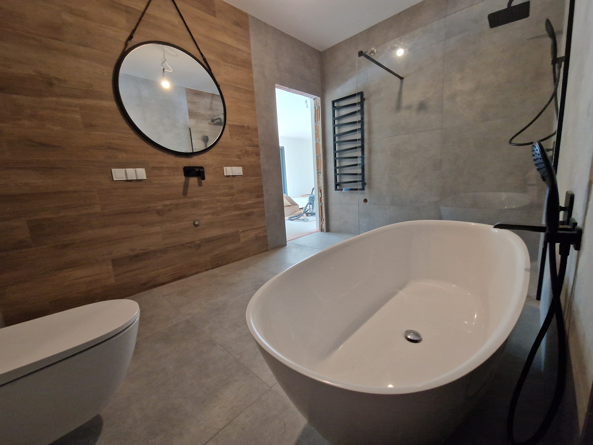 Nowoczesna łazienka z wolnostojącą białą wanną, drewnopodobną okładziną ściany i okrągłym lustrem na czarnym pasku, widoczny grzejnik drabinkowy i czarna armatura.