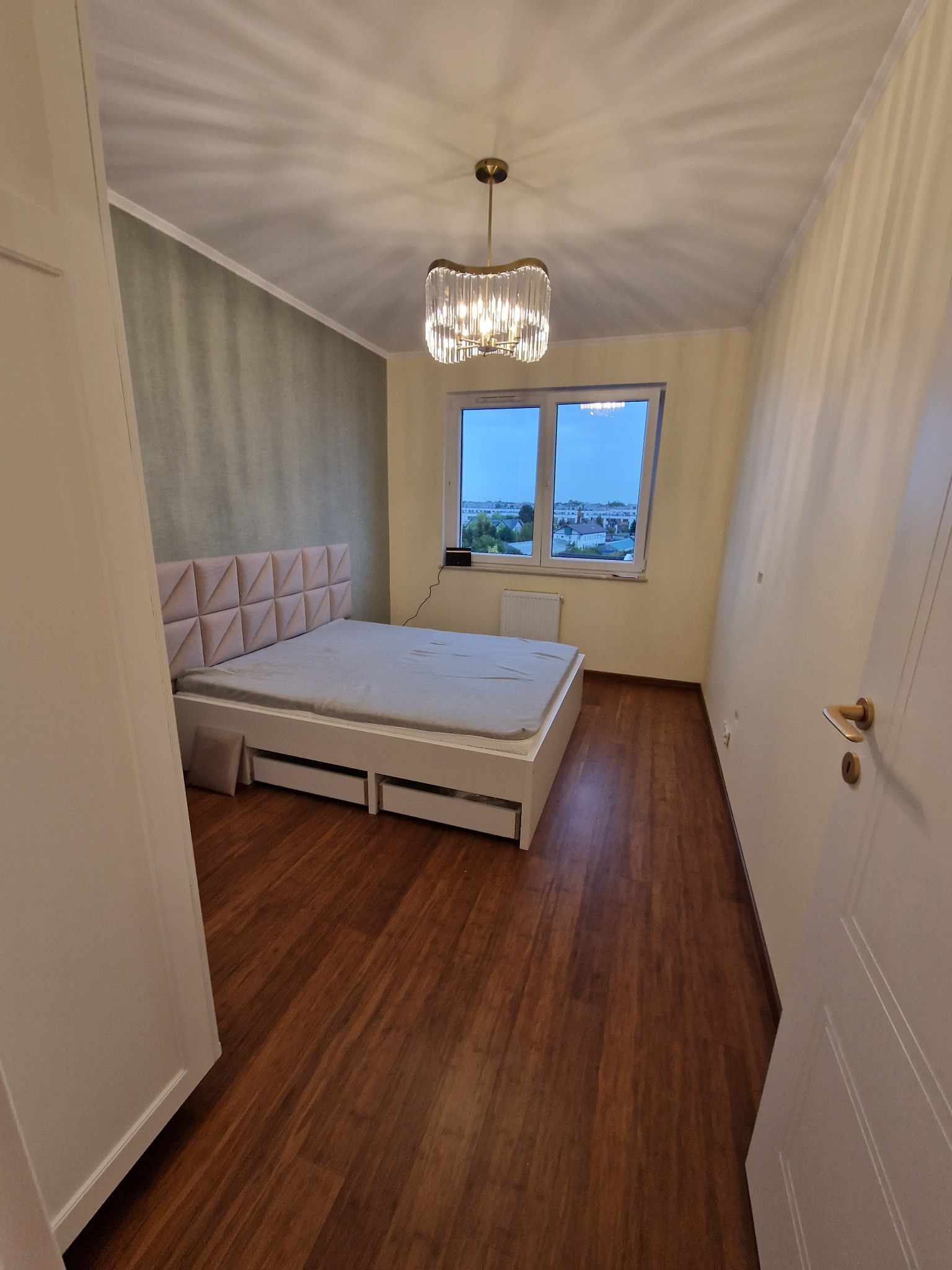 Nowoczesna sypialnia z łóżkiem z pikowanym zagłówkiem, drewnianą podłogą i ozdobną lampą sufitową, widok zza uchylonych drzwi.