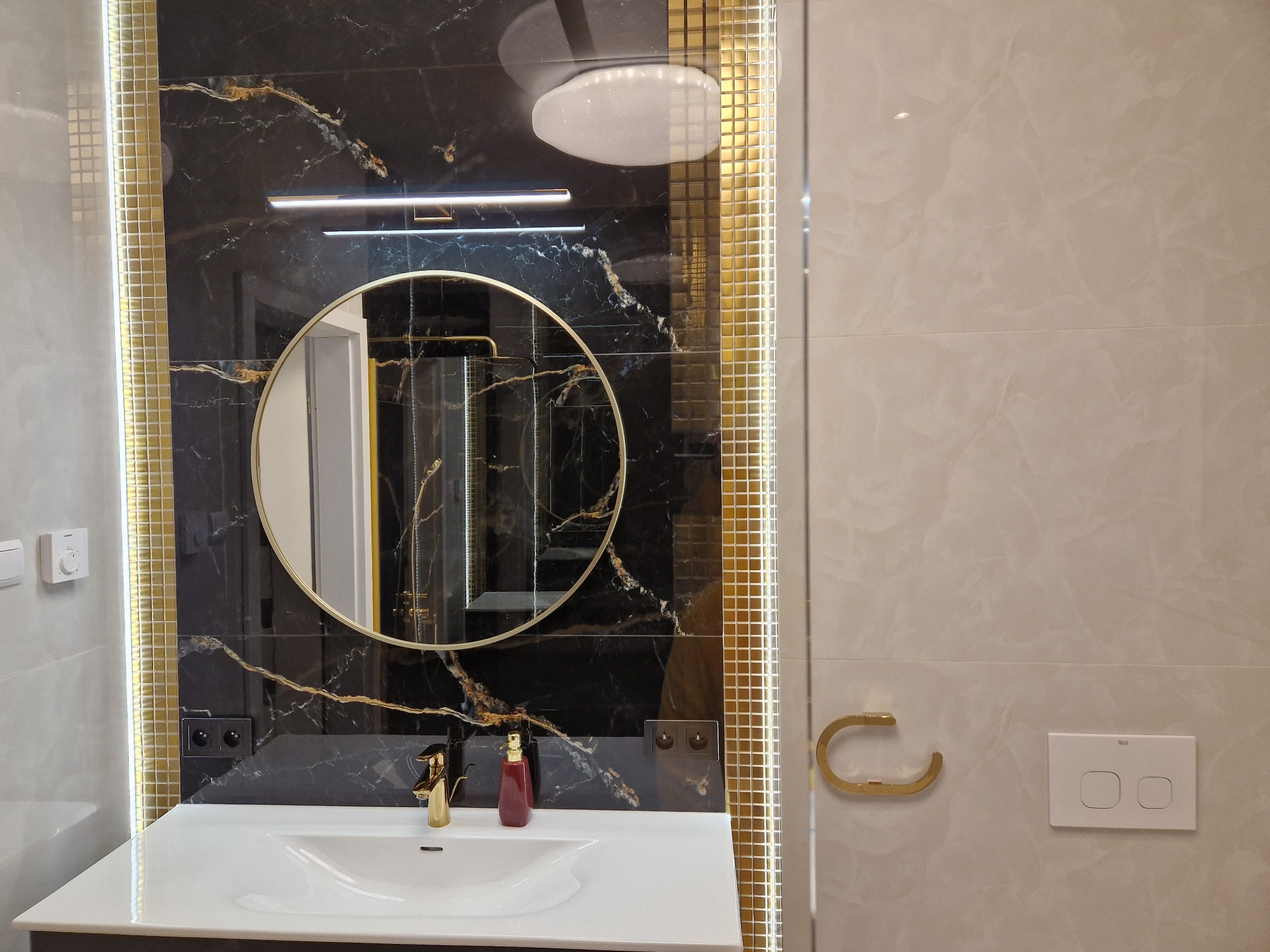 Elegancka łazienka: czarny marmur z żyłami złota, okrągłe lustro w złotej ramie, złota armatura i dozownik mydła, oświetlenie LED wzdłuż krawędzi płytek.