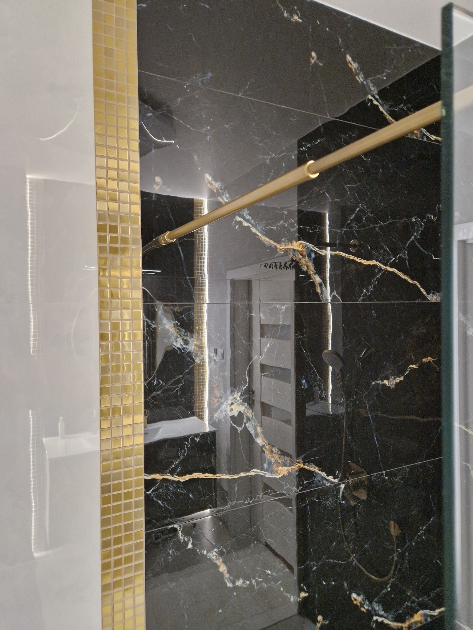 Wrocław: Detal wykończenia łazienki z czarną marmurową płytką z złotymi żyłami, obok biała ściana z pionowym pasem złotych mozaikowych płytek, widoczny fragment kabiny prysznicowej.