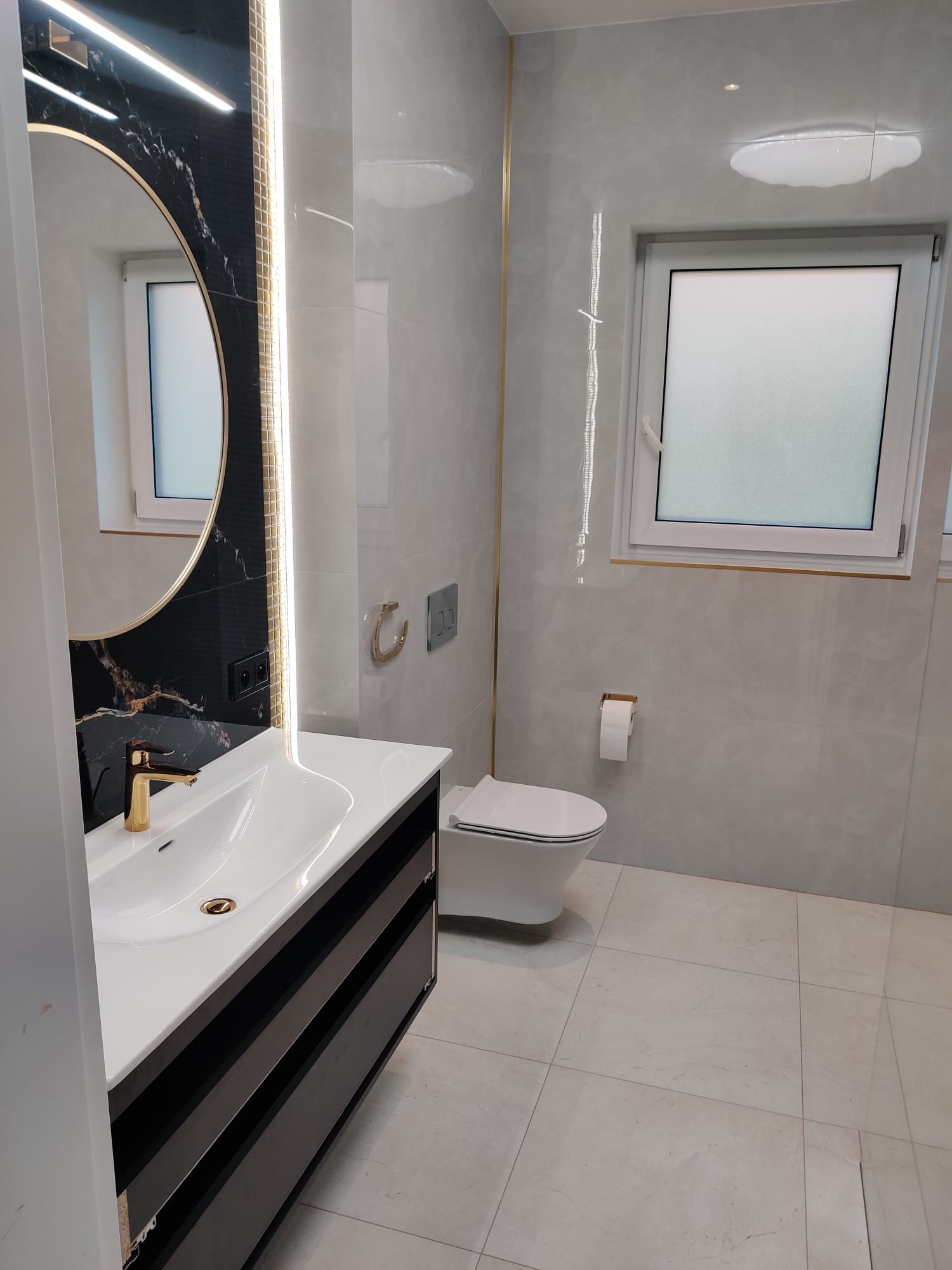 Nowoczesna łazienka z marmurowymi płytkami, owalnym lustrem z podświetleniem LED, złotą armaturą i wiszącą toaletą, oświetlona punktowo.