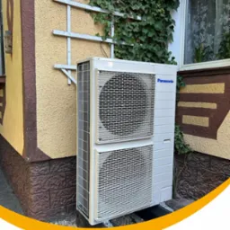 Zewnętrzna jednostka klimatyzacji Panasonic zamontowana na elewacji budynku z dekoracyjną kratką i pnącą roślinnością.