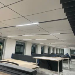 Wnętrze biura w trakcie montażu: widoczne sufitowe panele akustyczne w odcieniach szarości, biurka z regulowaną wysokością, częściowo zabezpieczone folią, oraz elementy instalacji elektrycznej...