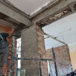 DreamBuilder - Pracownik na rusztowaniu wzmacnia strop z odsłoniętą cegłą metalowymi profilami mocowanymi na śruby, butelka wody na podeście rusztowania.