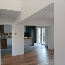 DreamBuilder - Wnętrze wyremontowanego mieszkania z drewnianą podłogą, białymi ścianami i widokiem na kuchnię z wyspą i czarną lodówką.