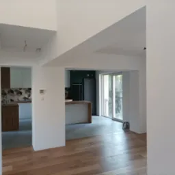 Wnętrze wyremontowanego mieszkania z drewnianą podłogą, białymi ścianami i widokiem na kuchnię z wyspą i czarną lodówką.