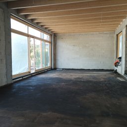 DreamBuilder - Surowe wnętrze domu w trakcie budowy, z surowymi ścianami z białego betonu komórkowego, drewnianym stropem i wylaną, ciemną posadzką. Duże okna przesuwne z widokiem na zieleń na zewnątrz.