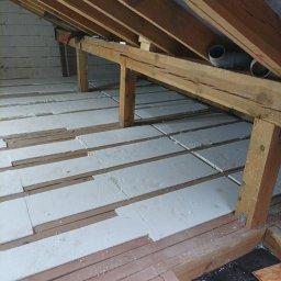 DreamBuilder - Białe płyty izolacyjne układane między drewnianymi belkami stropu poddasza, widoczne elementy konstrukcji dachu i fragment ściany z bloczków.