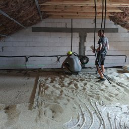 DreamBuilder - Prace na poddaszu: dwóch mężczyzn instaluje ogrzewanie podłogowe na warstwie izolacyjnej, widoczne worki z materiałem izolacyjnym i ocieplone skosy dachu.