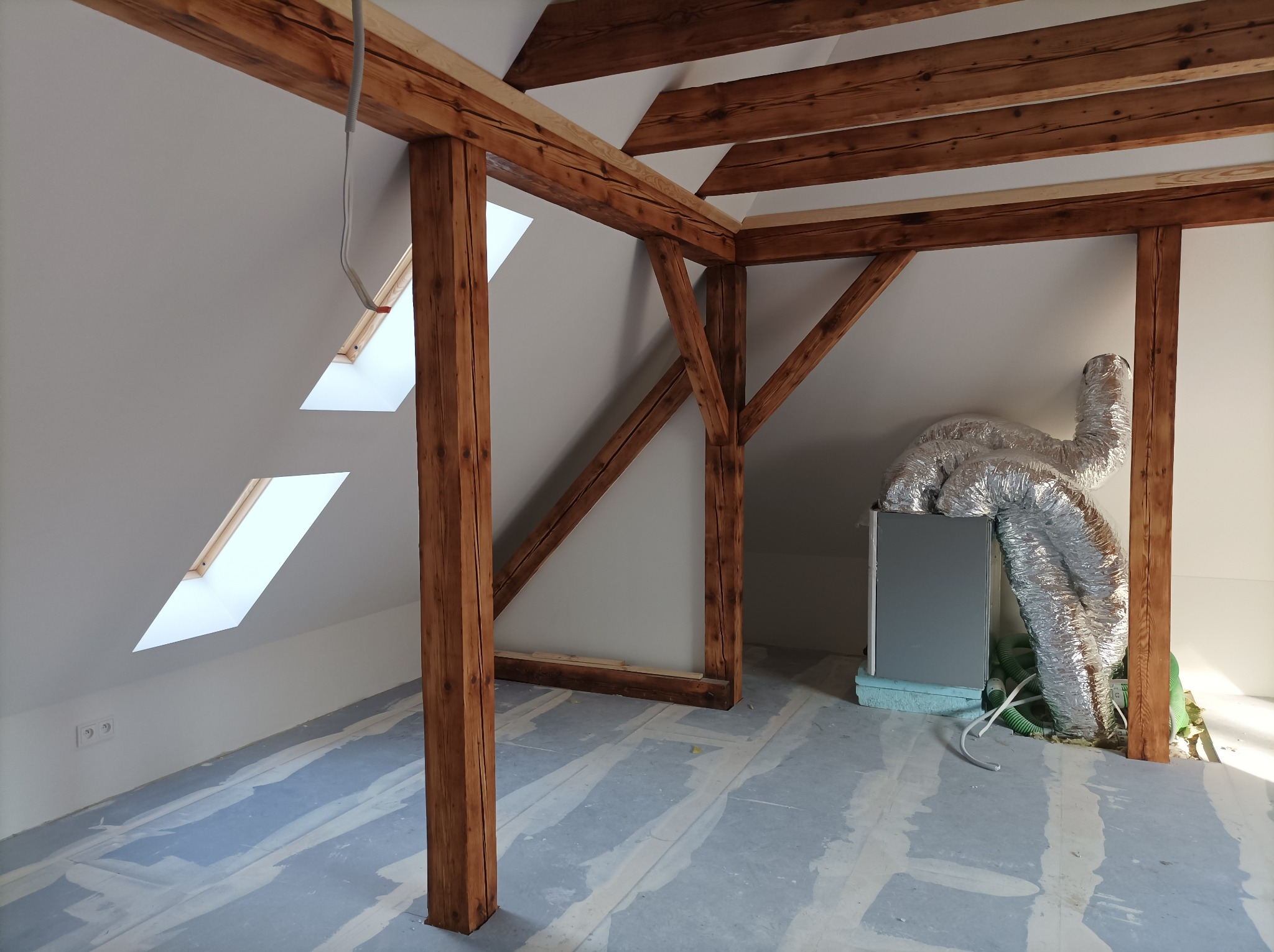 Poddasze z drewnianą konstrukcją więźby dachowej, dwoma oknami dachowymi i systemem wentylacji z metalową rurą podłączoną do urządzenia na podłodze.