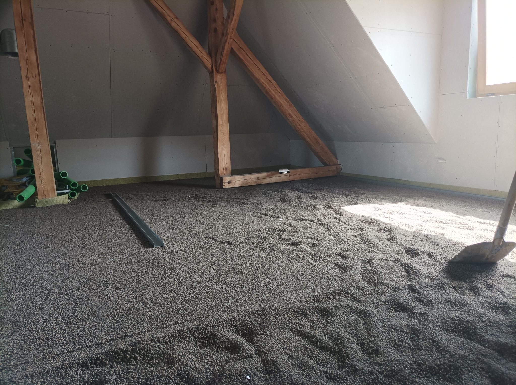 Poddasze w trakcie izolacji: widoczne drewniane elementy konstrukcyjne, płyty kartonowo-gipsowe na ścianach i podłodze pokrytej granulatem izolacyjnym, z rurami instalacyjnymi w rogu i łopatą...