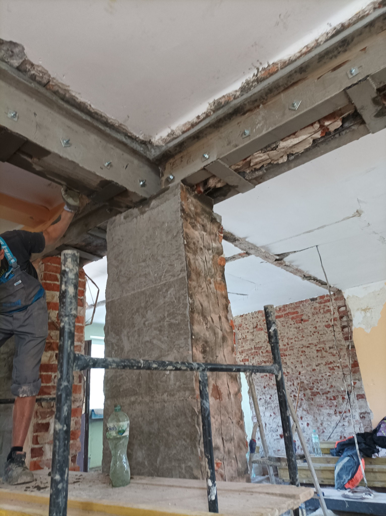 Pracownik na rusztowaniu wzmacnia strop z odsłoniętą cegłą metalowymi profilami mocowanymi na śruby, butelka wody na podeście rusztowania.