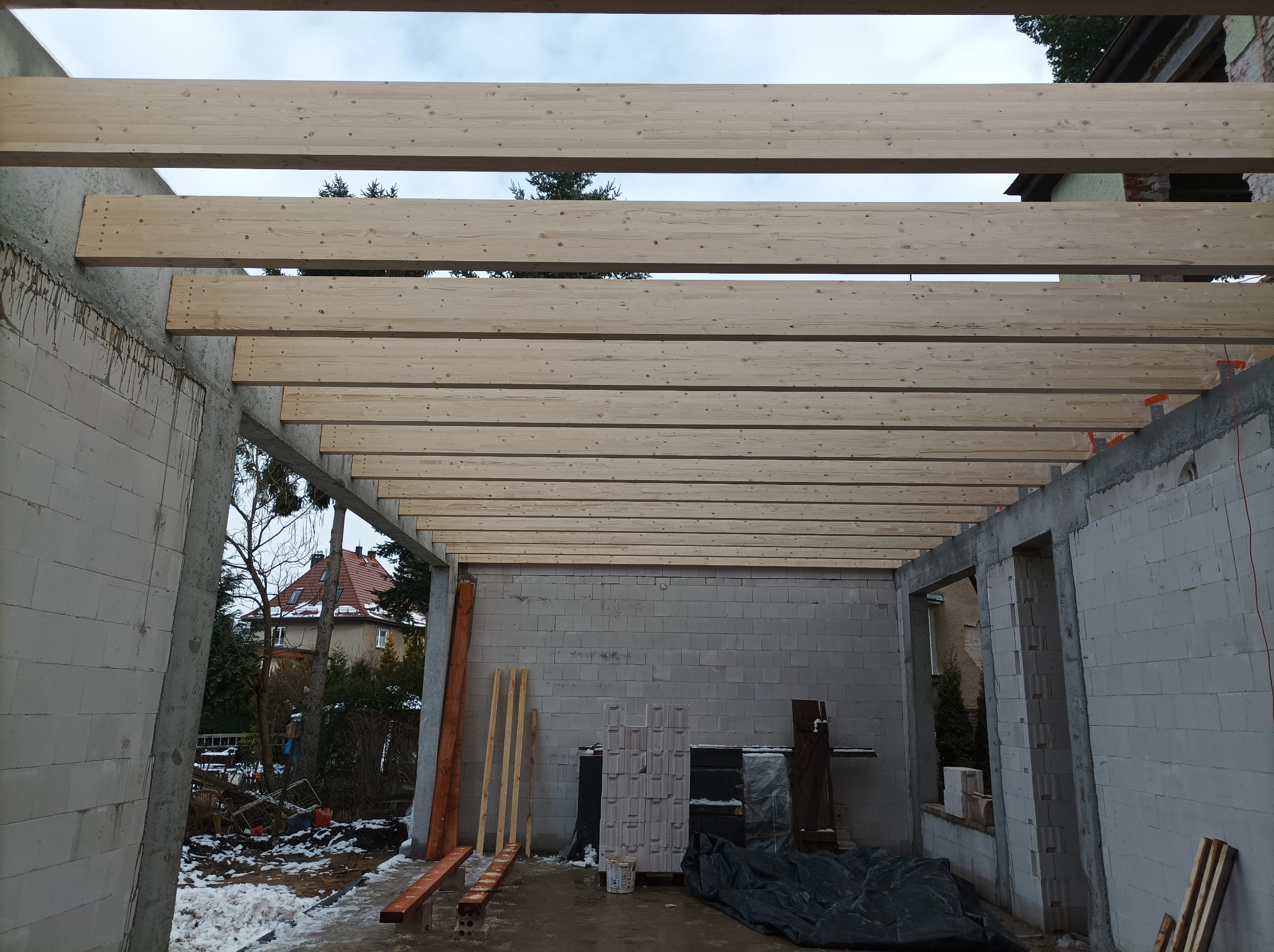 Surowy stan budowy z widoczną drewnianą konstrukcją dachu, bloczki betonowe, materiały budowlane na ziemi i dom w tle.