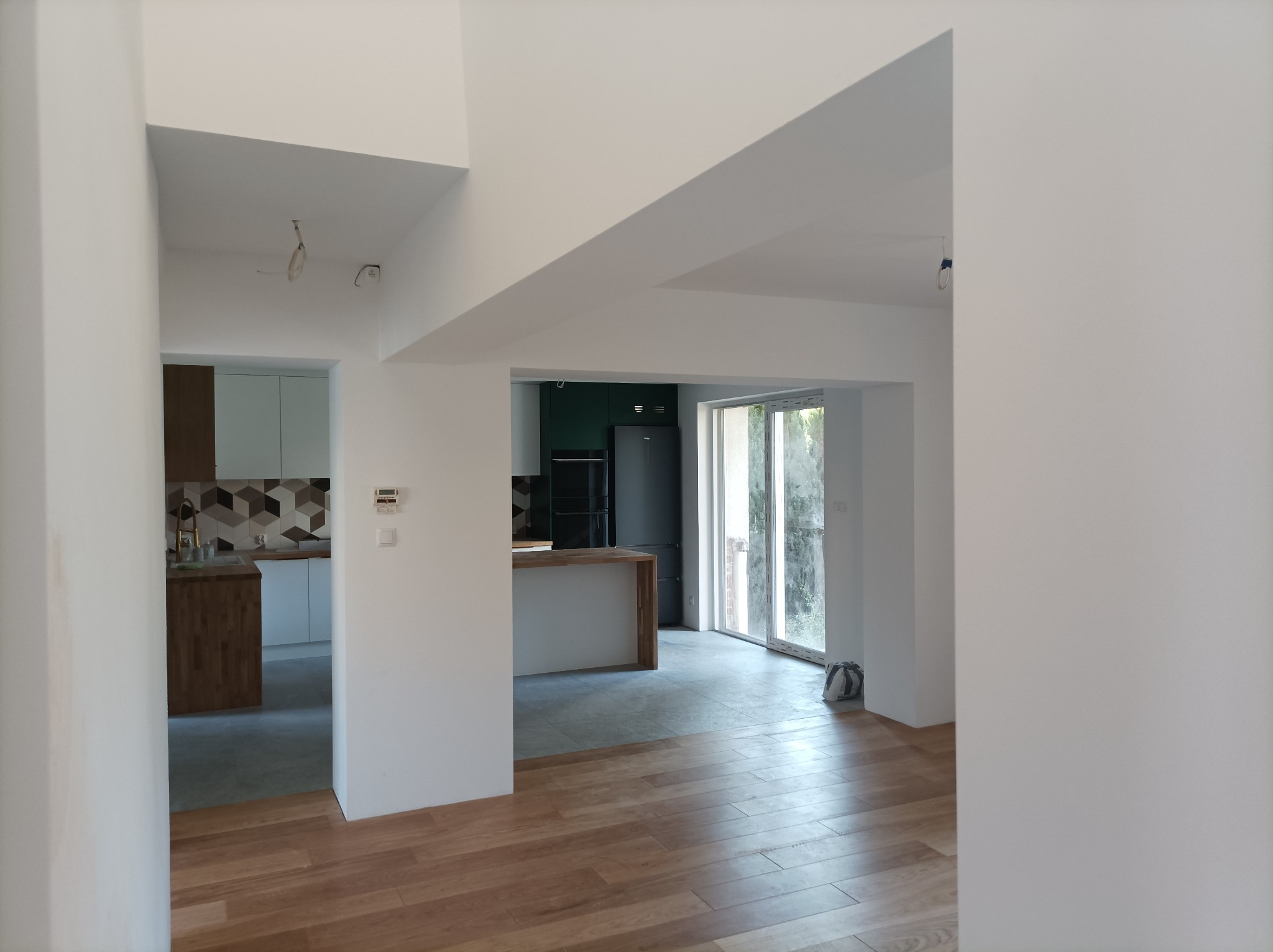 Wnętrze wyremontowanego mieszkania z drewnianą podłogą, białymi ścianami i widokiem na kuchnię z wyspą i czarną lodówką.