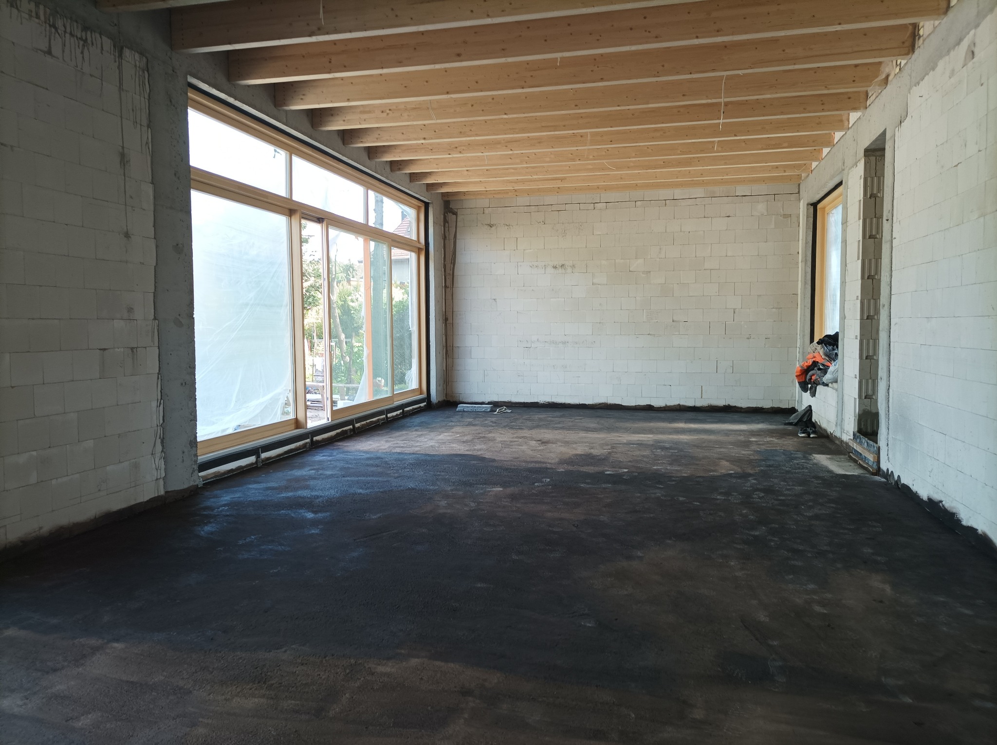 Surowe wnętrze domu w trakcie budowy, z surowymi ścianami z białego betonu komórkowego, drewnianym stropem i wylaną, ciemną posadzką. Duże okna przesuwne z widokiem na zieleń na zewnątrz.