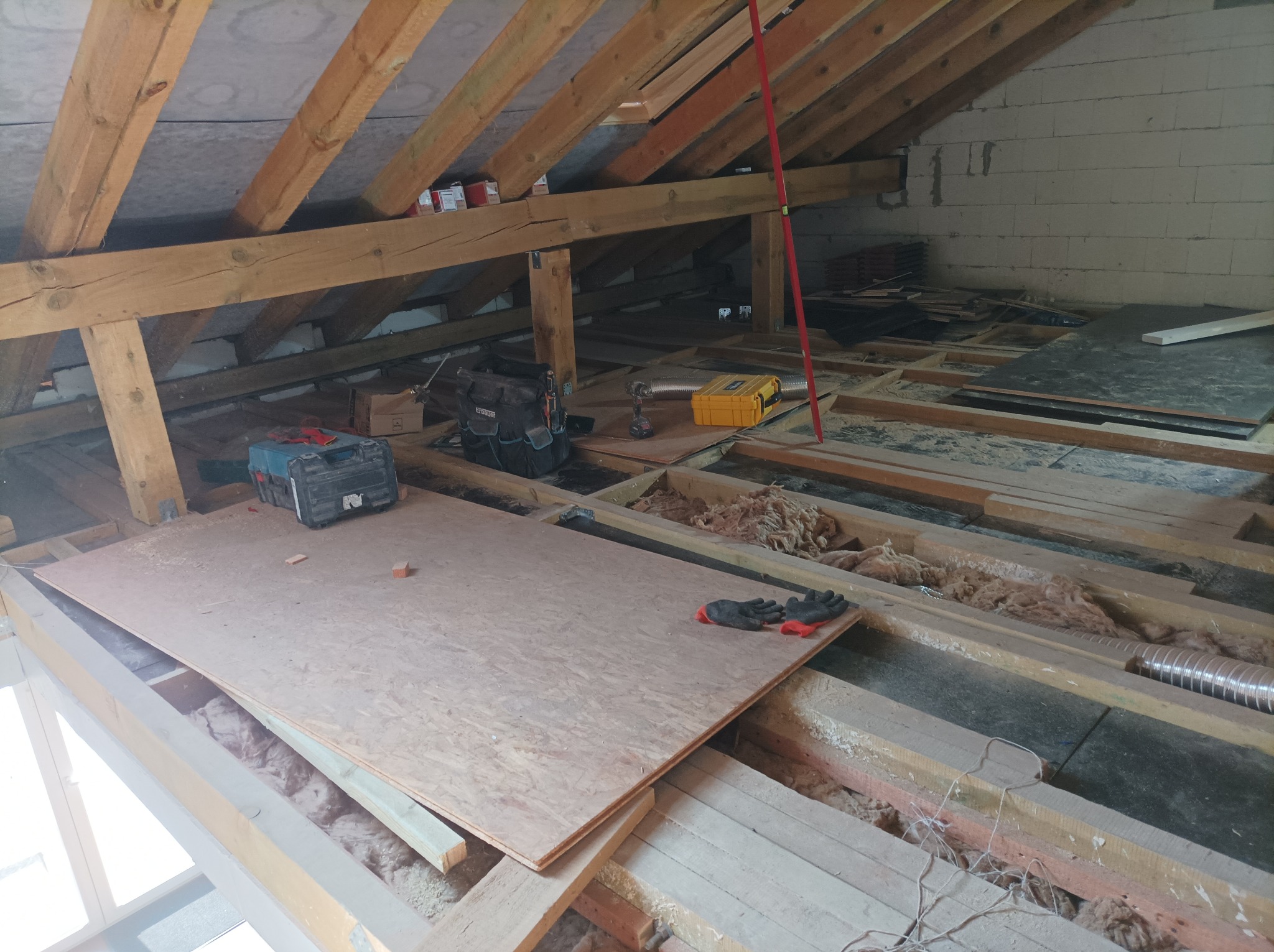 Poddasze w trakcie ocieplania z widoczną konstrukcją drewnianą, izolacją, narzędziami i płytą OSB na podłodze.