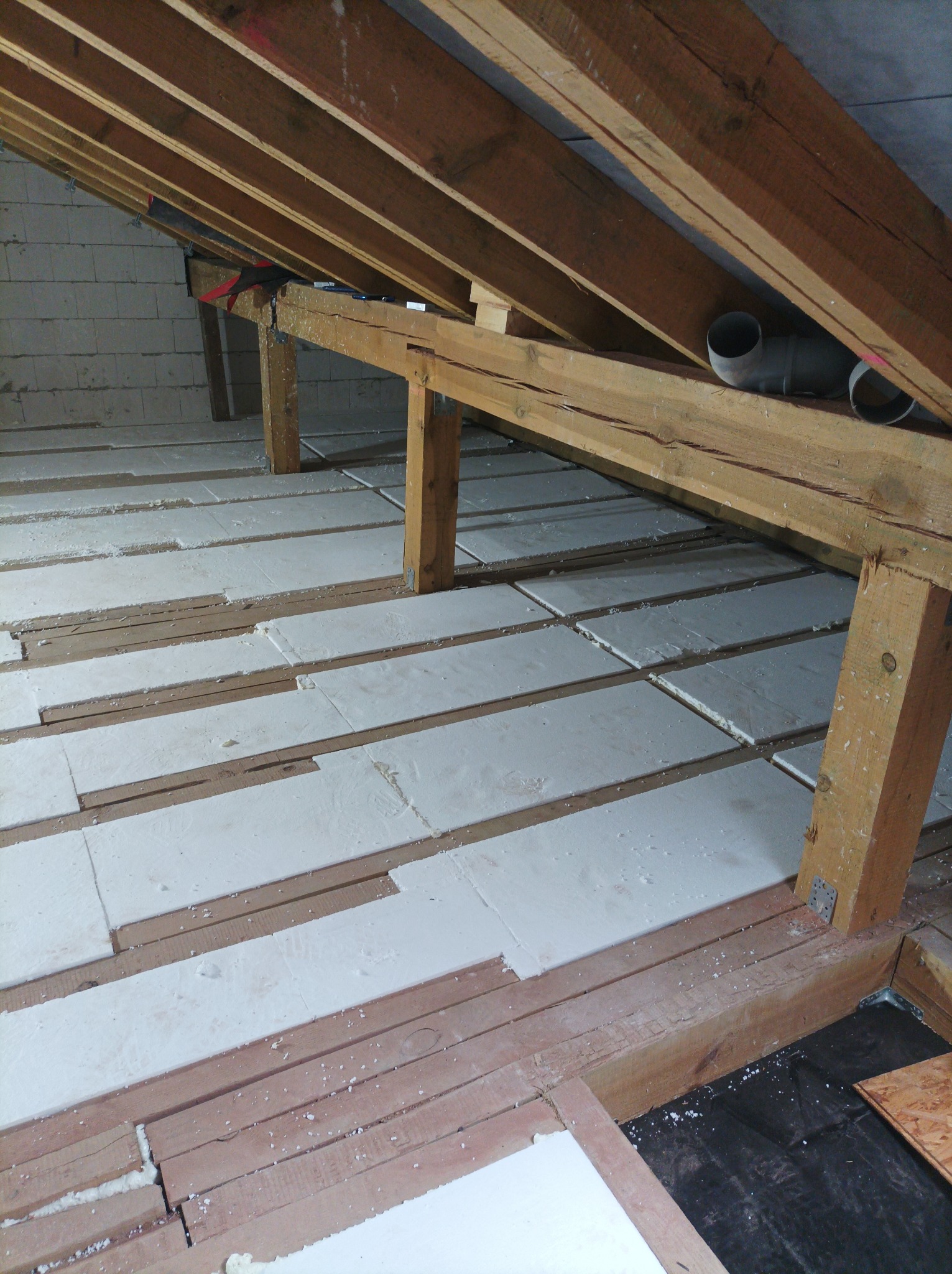 Białe płyty izolacyjne układane między drewnianymi belkami stropu poddasza, widoczne elementy konstrukcji dachu i fragment ściany z bloczków.