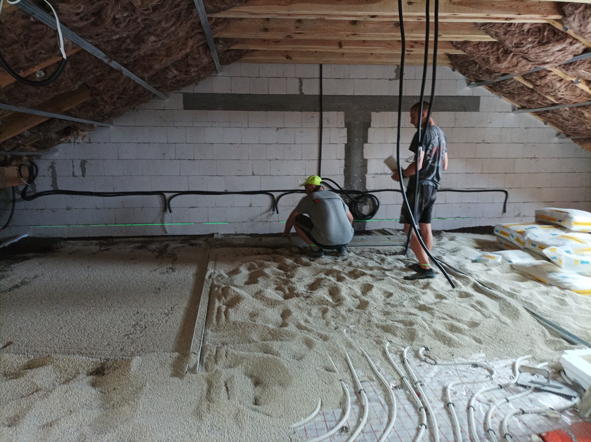 Prace na poddaszu: dwóch mężczyzn instaluje ogrzewanie podłogowe na warstwie izolacyjnej, widoczne worki z materiałem izolacyjnym i ocieplone skosy dachu.