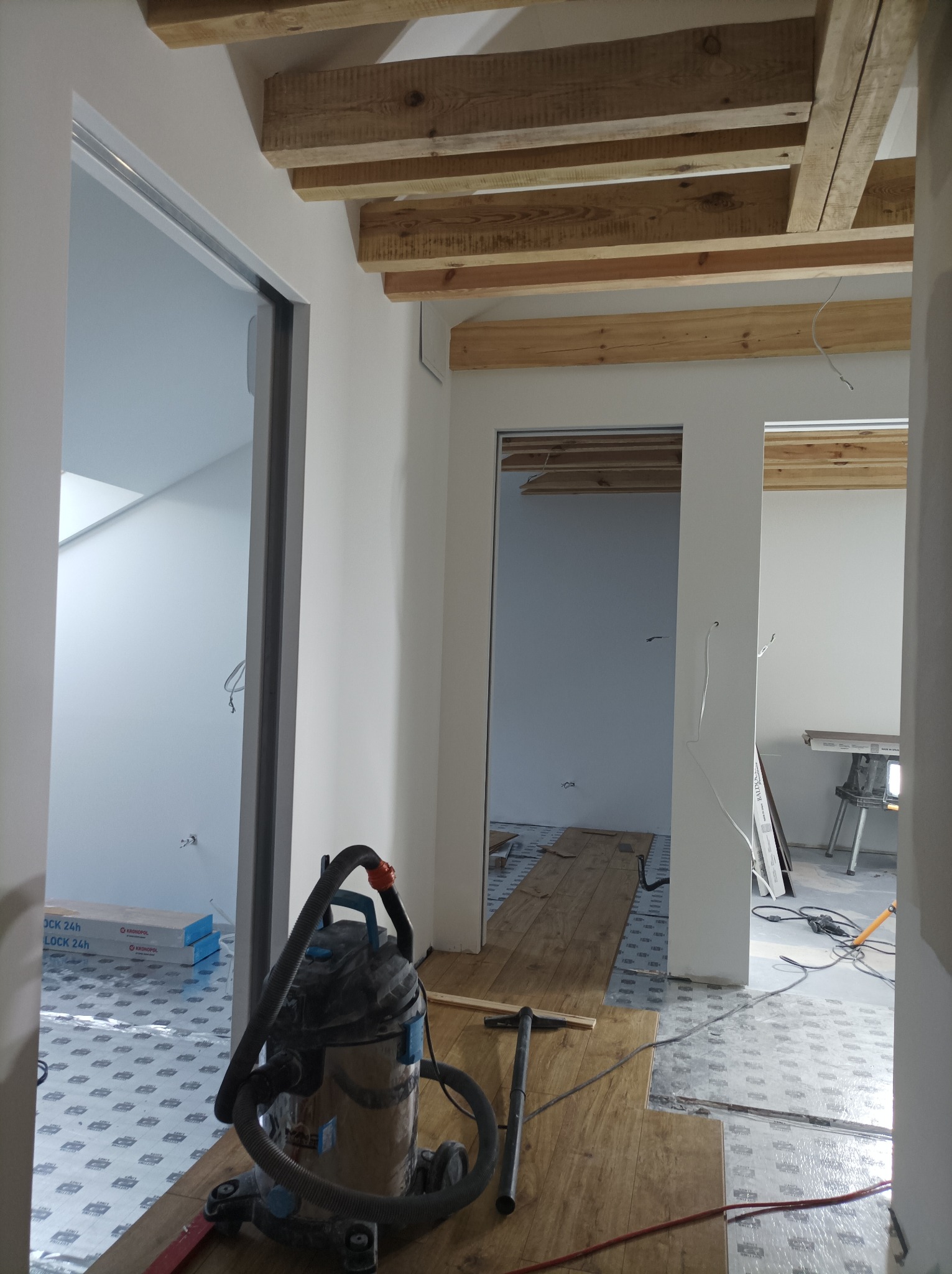Wnętrze w trakcie prac wykończeniowych: widoczne drewniane belki sufitowe, przygotowane podłoże pod panele, odkurzacz przemysłowy i narzędzia.