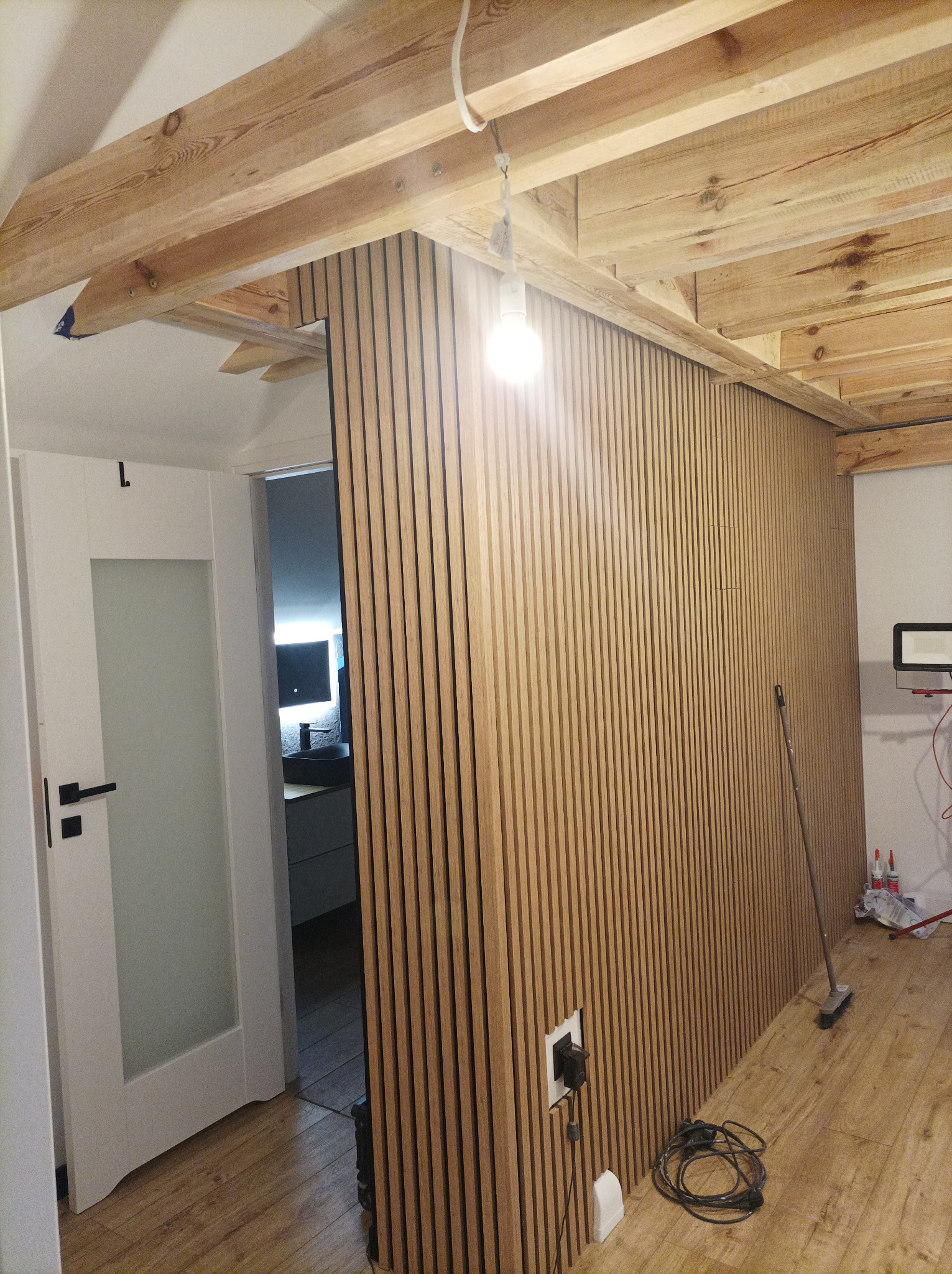 Drewniane lamele ścienne w jasnym odcieniu, montowane w pomieszczeniu z drewnianą konstrukcją stropu, widoczny fragment białych drzwi i podłoga z paneli, gniazdko elektryczne w ścianie, kabel...