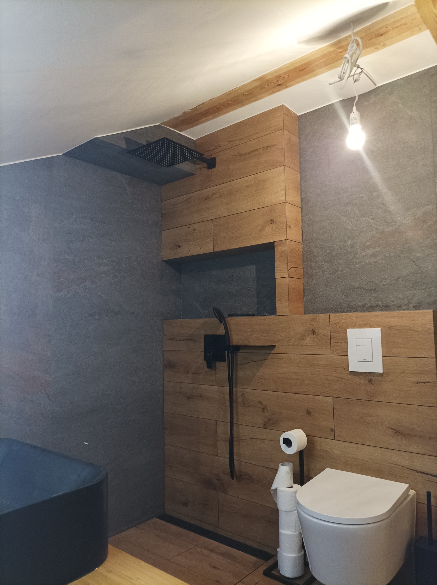 Nowoczesna łazienka z drewnopodobnymi panelami ściennymi, czarną armaturą prysznicową i wiszącą toaletą. Widoczna surowa żarówka i drewniany element konstrukcyjny na suficie.