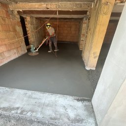 Kachel Bau sp. z o.o. - Pracownik w surowym wnętrzu wygładza posadzkę jastrychową za pomocą zacieraczki mechanicznej. Widoczne ceglane ściany i betonowy strop. Przewody pod sufitem.