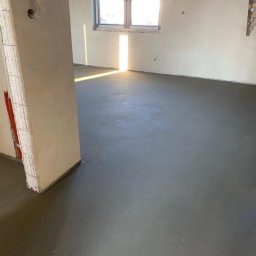 Kachel Bau sp. z o.o. - Gładka, szara posadzka jastrychowa w jasnym pomieszczeniu z oknem i widocznymi instalacjami w ścianie. Świeżo wylana podłoga w trakcie schnięcia.