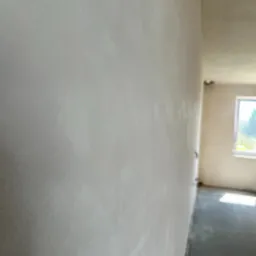 Ściana po nałożeniu tynku maszynowego, widok z boku, z delikatną fakturą, w tle widoczne okno i surowy strop. Oświetlenie naturalne.