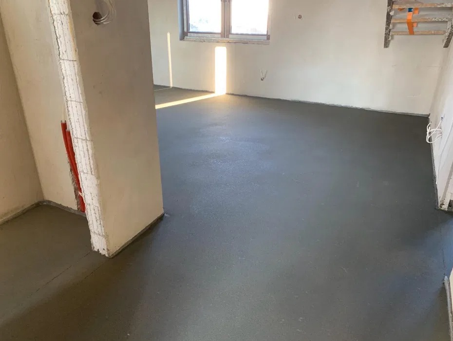 Gładka, szara posadzka jastrychowa w jasnym pomieszczeniu z oknem i widocznymi instalacjami w ścianie. Świeżo wylana podłoga w trakcie schnięcia.