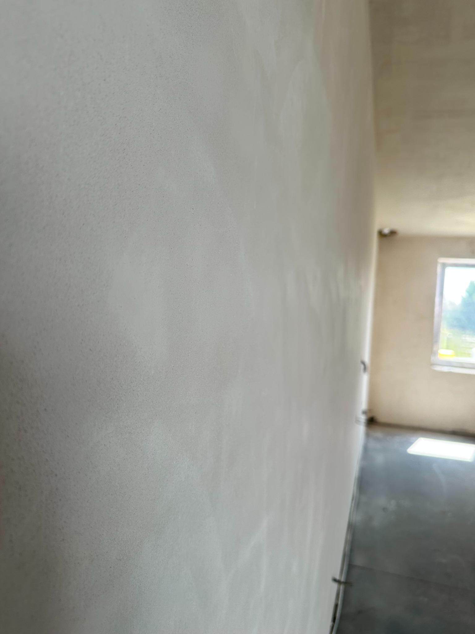 Ściana po nałożeniu tynku maszynowego, widok z boku, z delikatną fakturą, w tle widoczne okno i surowy strop. Oświetlenie naturalne.