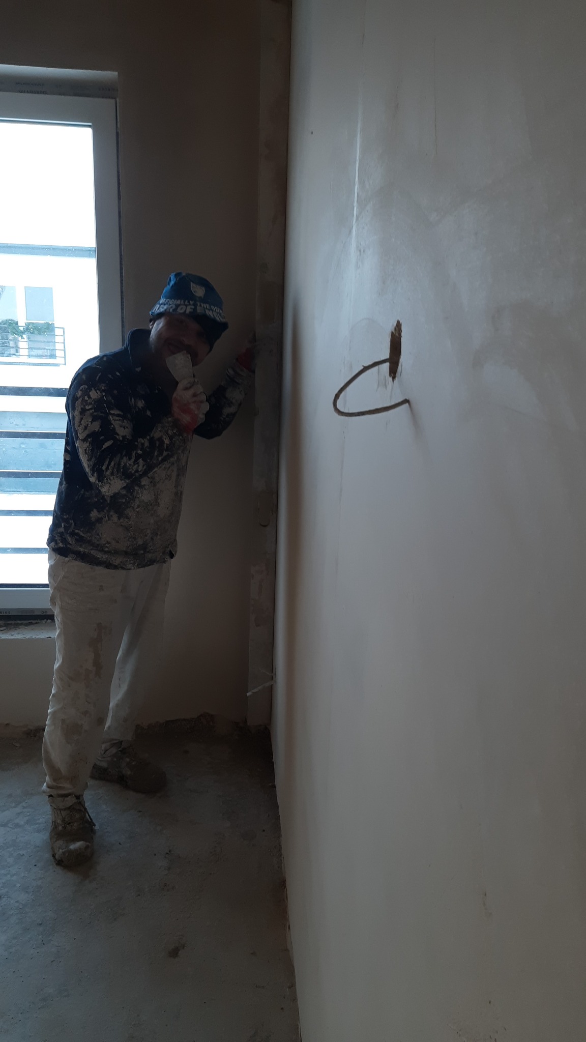 Pracownik w ubrudzonym stroju roboczym wygładza ścianę szpachelką, widoczny wystający kabel elektryczny ze ściany.