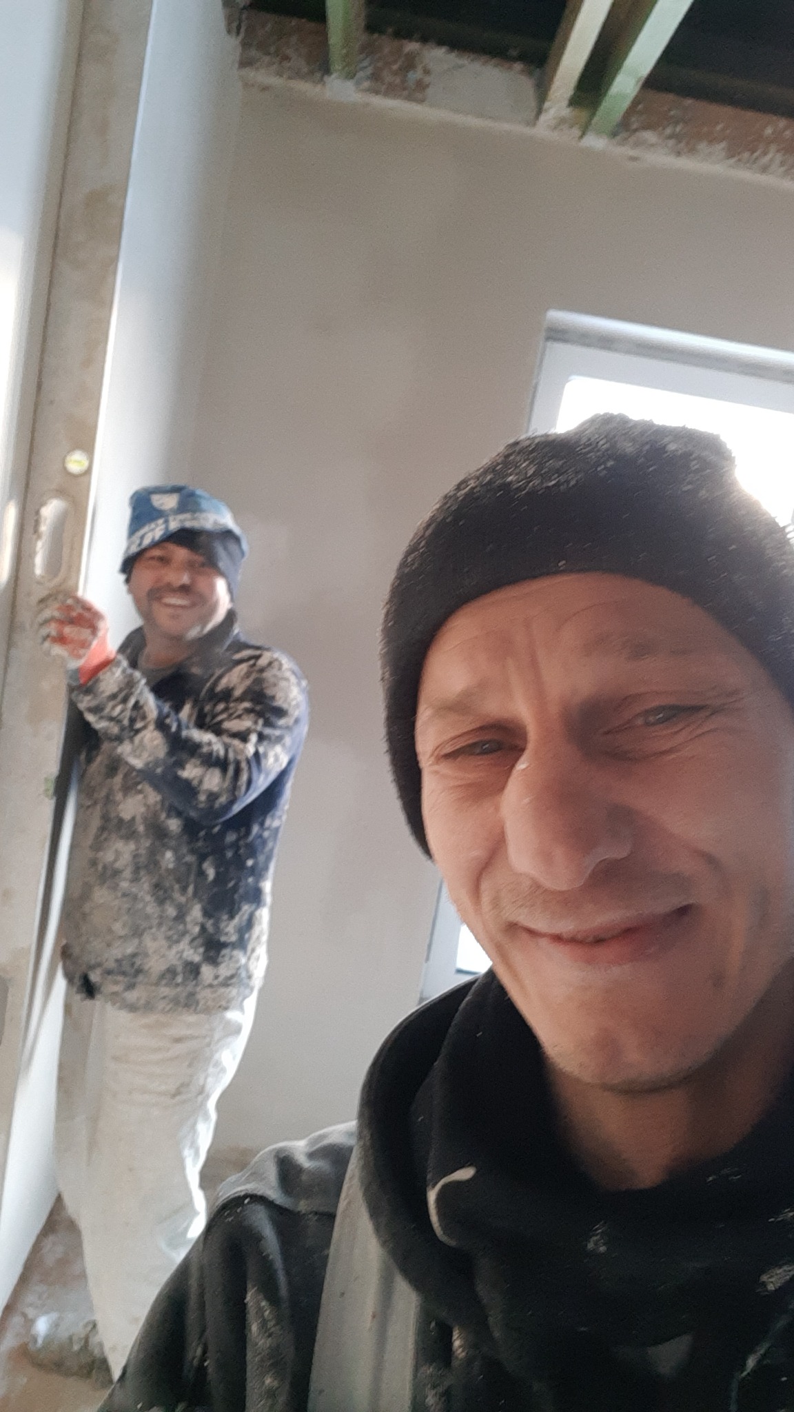 Dwóch mężczyzn w trakcie prac wykończeniowych wnętrza; jeden z poziomicą przy ścianie, obaj ubrani w robocze ubrania.