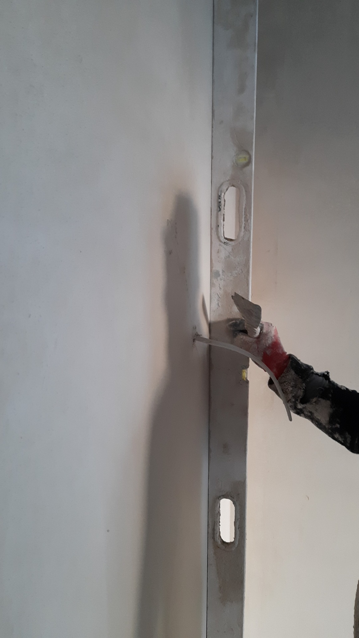 Praca z poziomnicą przy ścianie, ręka w rękawicy z zabrudzeniami, szpachelka przy otworze w ścianie.