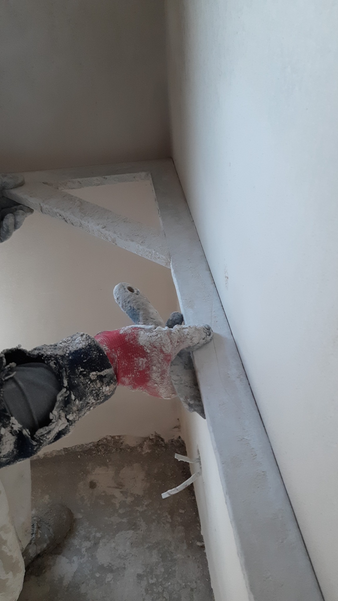 Pracownik w czerwonej rękawicy wyrównuje narożnik ściany za pomocą szpachelki, widoczne ślady zaprawy na ścianie, podłodze i ubraniu roboczym.