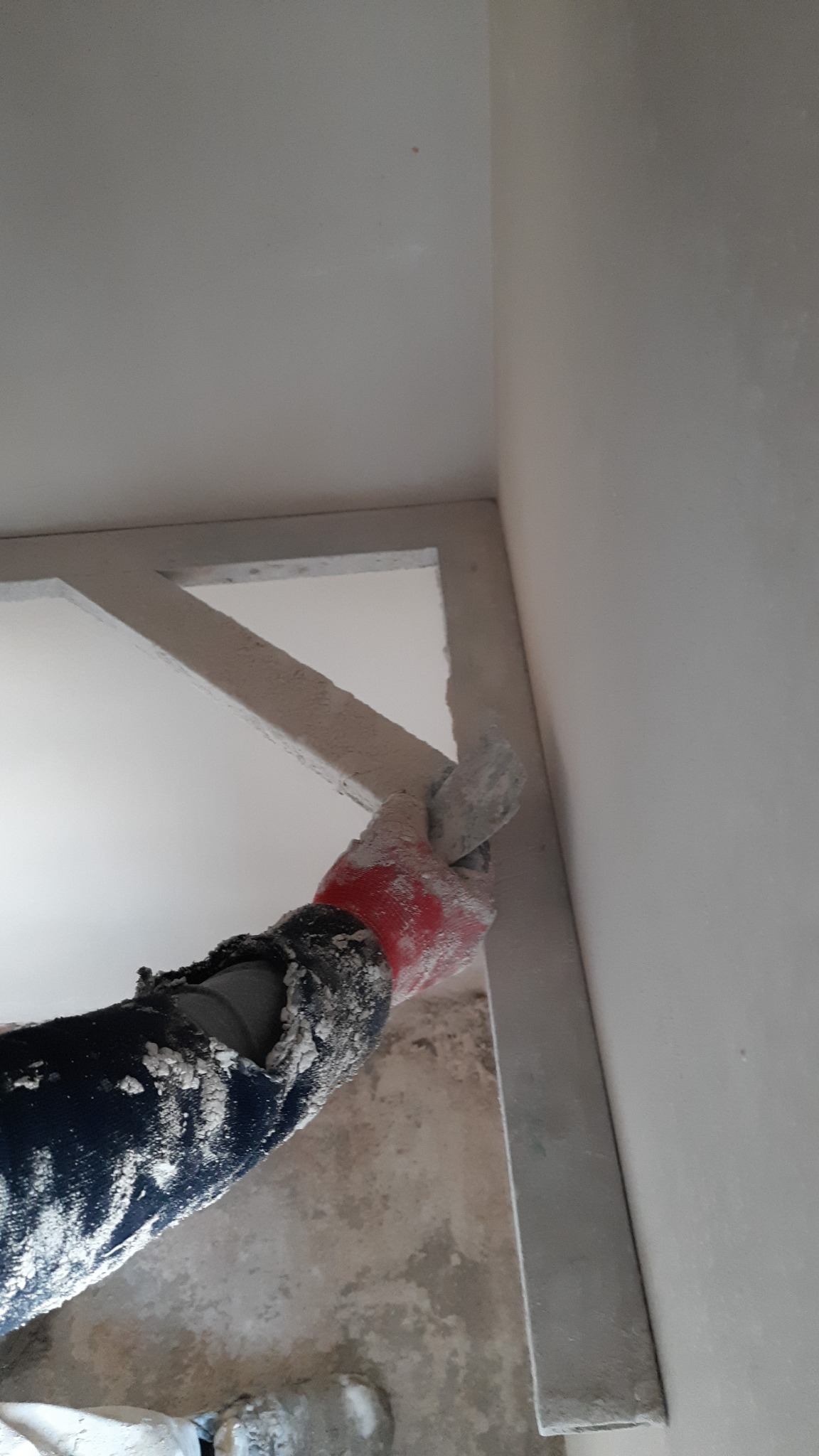 Praca z gładzią przy narożniku sufitu, widoczna ręka w czerwonej rękawicy i ubrudzona zaprawą odzież robocza.