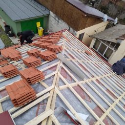 Burcea dominic - Montaż pomarańczowej dachówki na drewnianej konstrukcji dachu pokrytej membraną, widok z góry na budynki w tle.