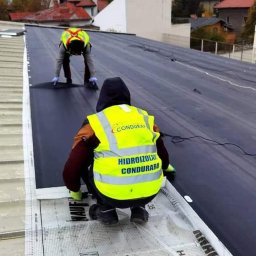 Burcea dominic - Dwóch pracowników w odblaskowych kamizelkach układa hydroizolację na płaskim dachu, widoczne rolki materiału i zgrzewarka.