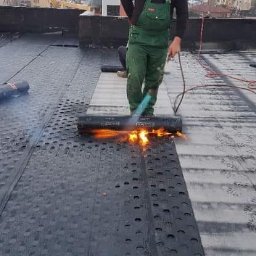 Burcea dominic - Pracownik w zielonym kombinezonie z palnikiem gazowym podczas układania papy termozgrzewalnej na dachu, widoczna butla gazowa i zgrzana powierzchnia dachu.