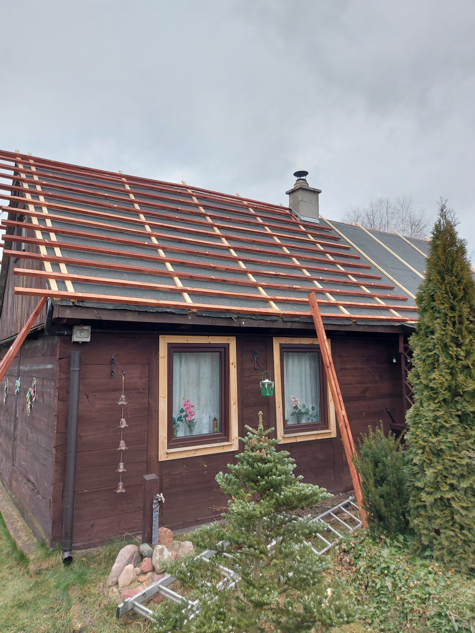 Dom z częściowo zdjętym poszyciem dachowym, widoczne drewniane łaty przygotowane pod nowe pokrycie, drabina oparta o ścianę, pochmurne niebo.