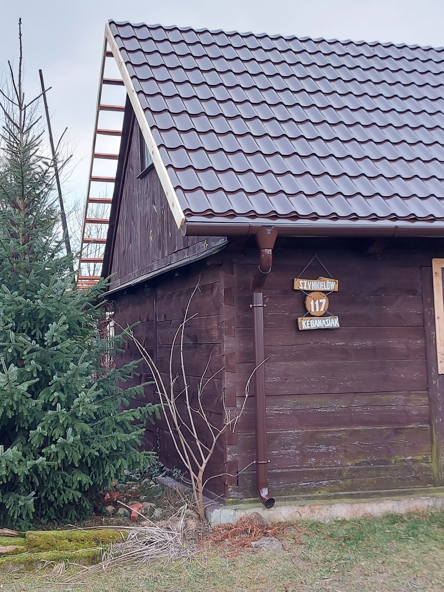 Brązowy dach z blachodachówki na drewnianym domu z brązowymi rynnami, widoczna drewniana konstrukcja dachu i tabliczka adresowa.