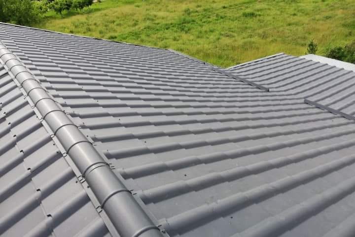 Szary dach z dachówki ceramicznej, widok z góry, w tle zielone drzewa i trawa.