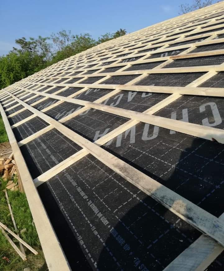 Konstrukcja dachu z membraną hydroizolacyjną i łatami, widok z dołu pod kątem, widoczny fragment nieba i drzew w tle. Na membranie widoczne napisy informacyjne.